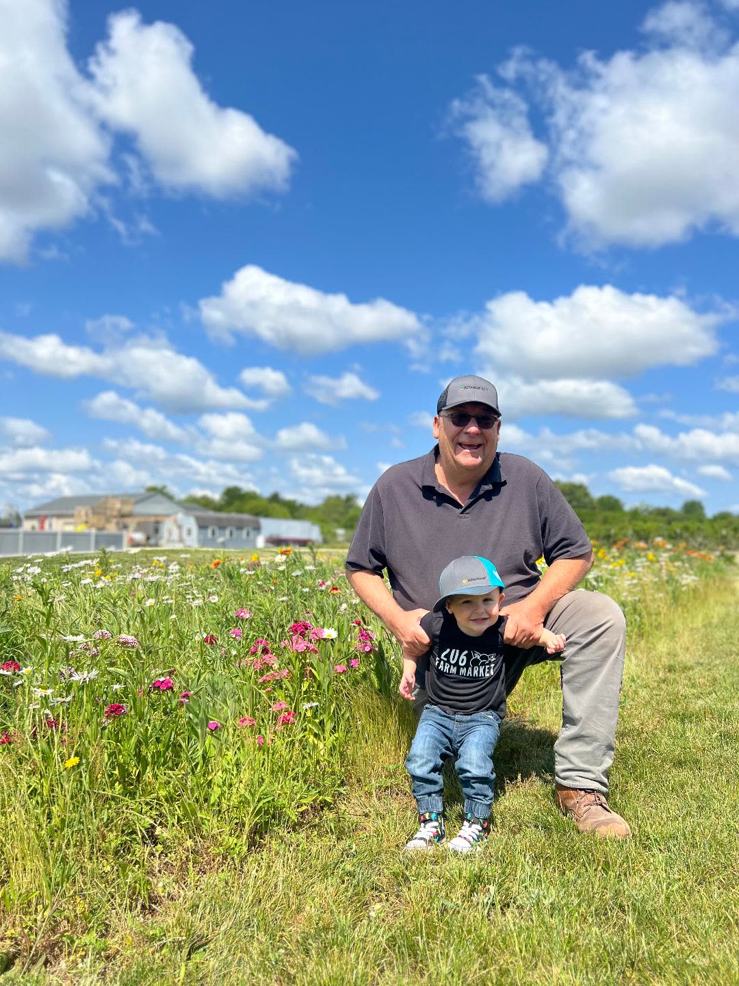PopPop kneeling in the wildflower field with G, who wears a 206 Farm Market shirt